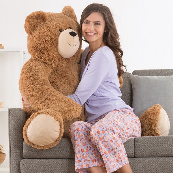 4' Big Hunka Love Bear - Seated golden brown bear with a female model in lavender pajamas on a sofa