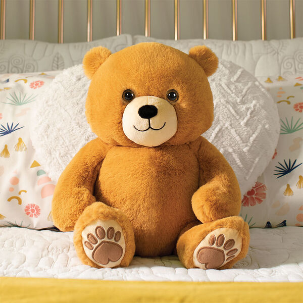 20" Hugsy the Teddy Bear - Front view of seated golden brown bear with light brown muzzle and embroidered foot pads in a bedroom scene