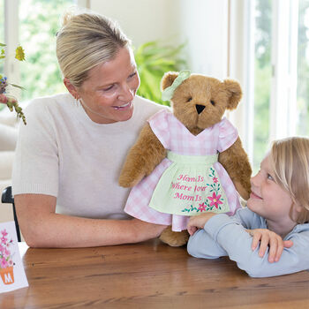 15" Home Is Where Your Mom Is Bear - Front view of standing jointed bear wearing a pink gingham dress, green bow and apron with floral embroidery and says "Home is Where Your Mom Is" with models